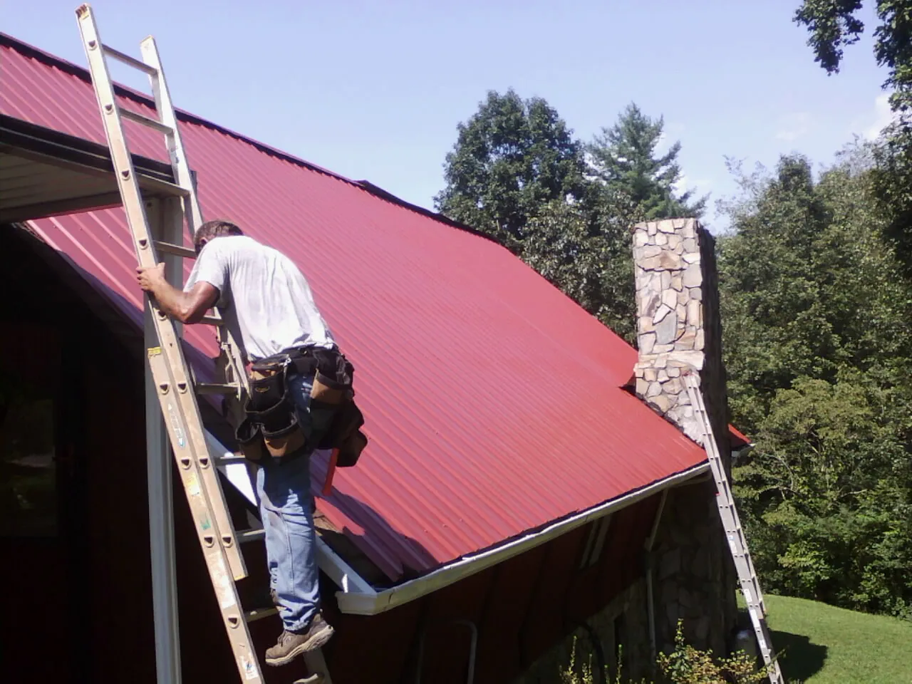 Roofer on a ladder beside a red metal roof and stone chimney — Soffit & Fascia Repair by Roofing Brothers in Wrentham